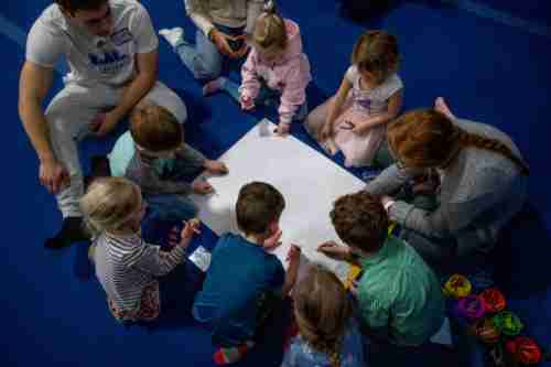 Two grand valley students and a group of preschool students lean over a white piece of poster board. There are cups of colorful coloring materials next to them.